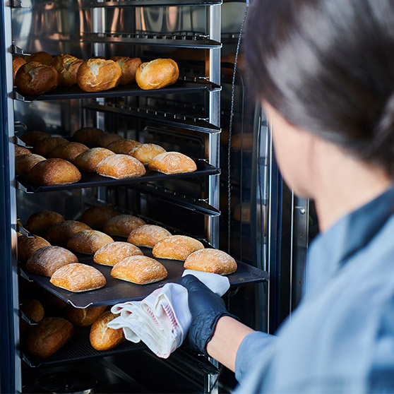 A person removes freshly baked bread rolls from Hobart's CHEF'S KOMBI using gloves and a cloth – multiple trays of golden-brown rolls in a professional oven.